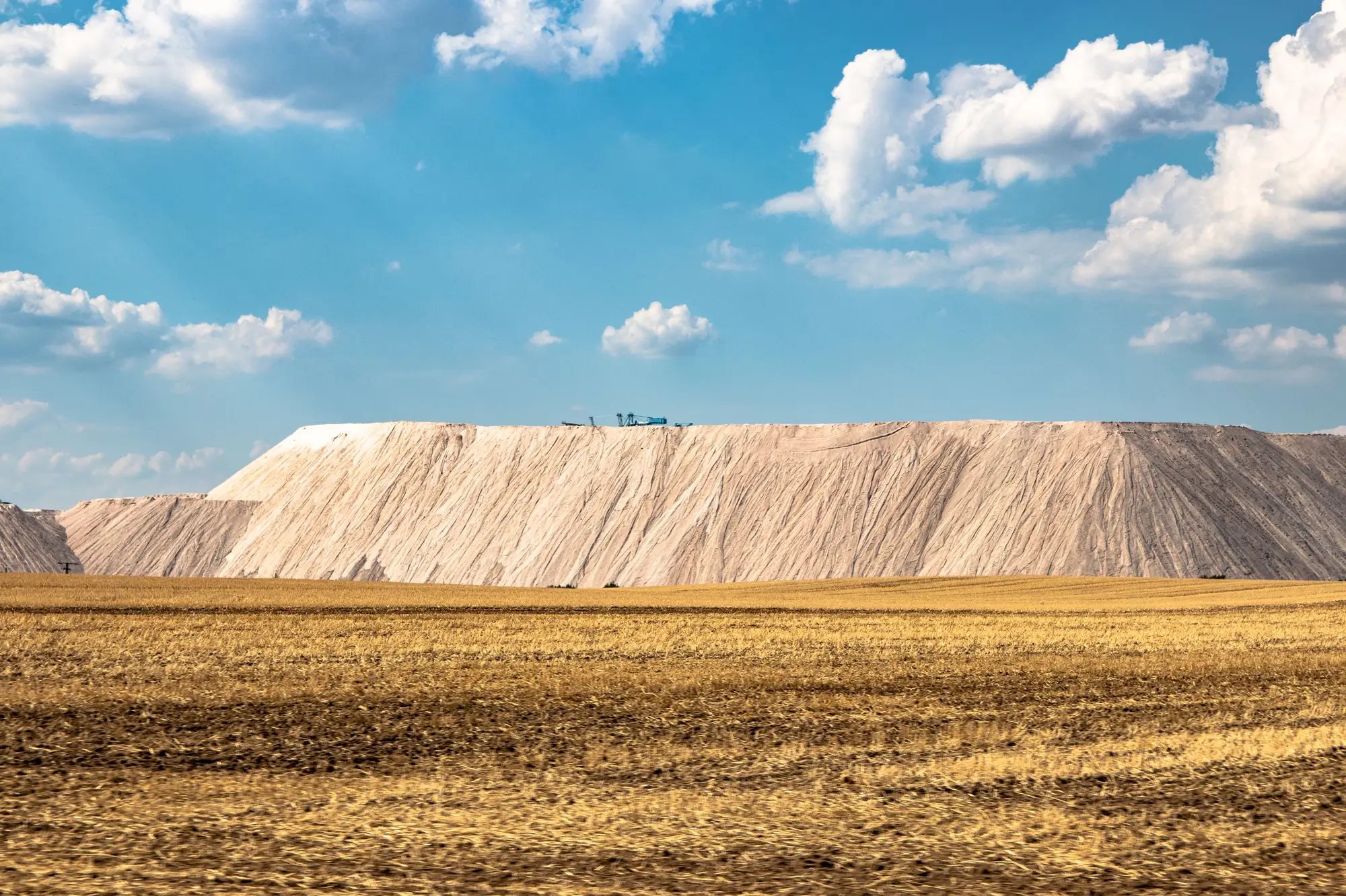 A pale spoil heap rising behind a harvested field under a blue sky
